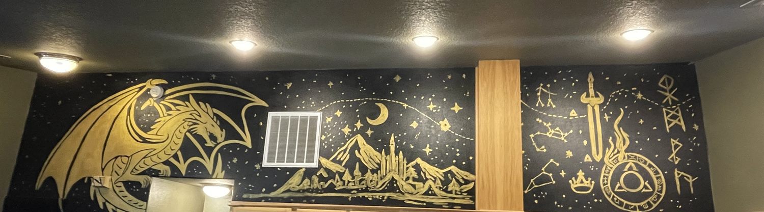 A black accent wall hand-painted in gold with a dragon, mountains and a castle, a crescent moon and stars, a sword wreathed in flame, constellations and runes, the mural that inspired the MOBS black-and-gold color scheme
